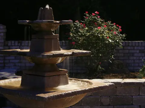 Tiered stone fountain lit at night against a brick wall with roses planted behind it