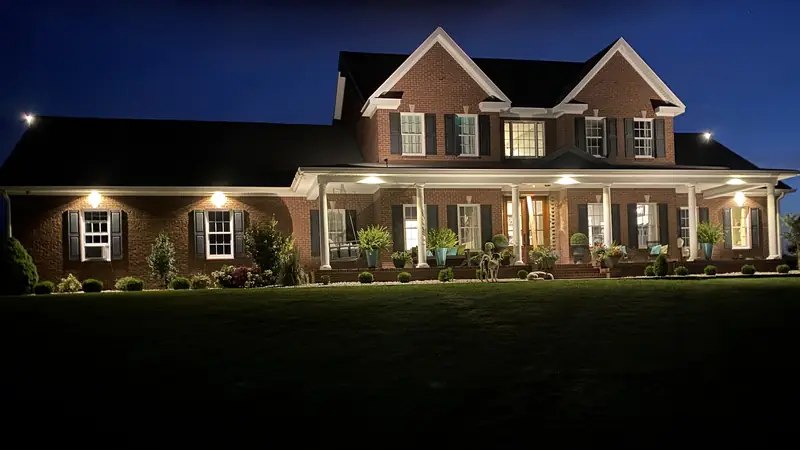 The same house with downlights in the porch ceiling and under the eaves and its shrubs lit