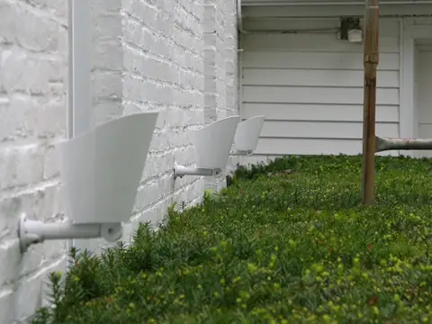 Painted brick wall in daylight with a pale fixture tucked against the base among ground cover