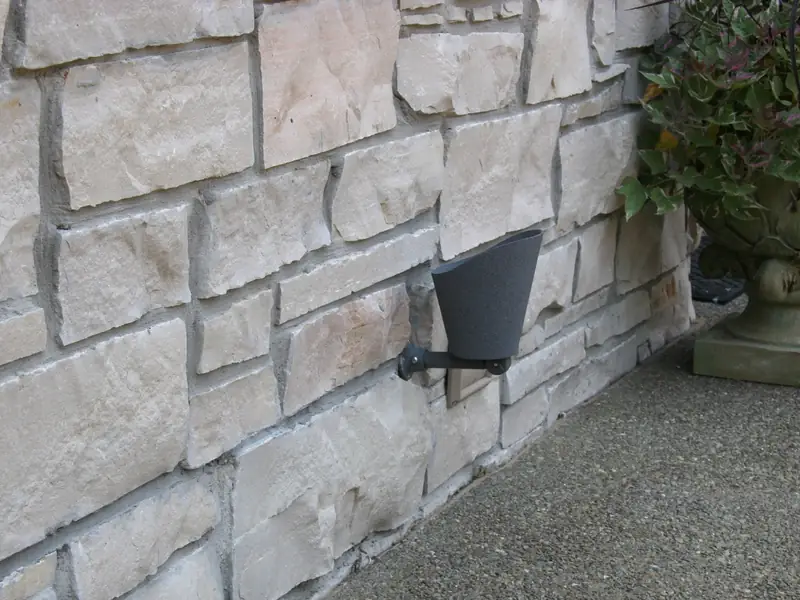 A small bronze fixture mounted flush against a stone wall, photographed in daylight and almost invisible against the coursing