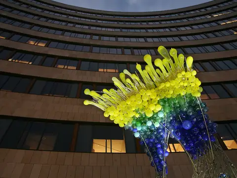 Large blown-glass sculpture lit in colour against a curved office building at dusk