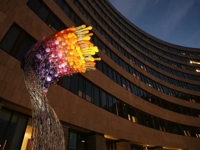 Tall blown-glass sculpture lit in warm and violet tones against a curved multi-storey building at dusk