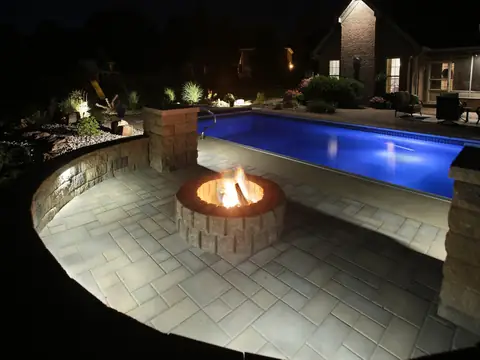Circular paver terrace with a fire pit beside a blue-lit pool
