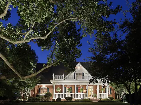 Uplit oak limbs framing a lit porch home at blue hour