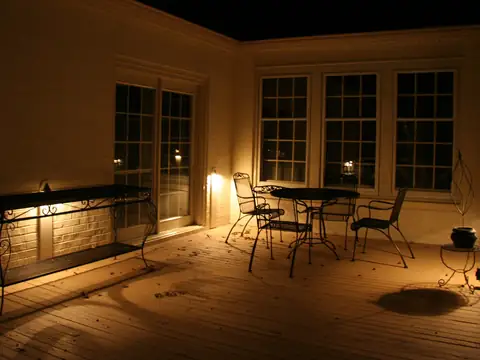 Timber deck at night with a table and chairs lit softly and the house windows glowing behind