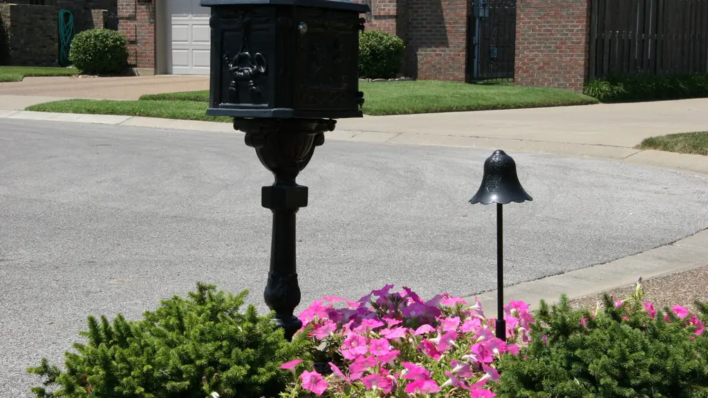 A black path light and a lamp post beside a driveway and flowering bed, photographed in daylight