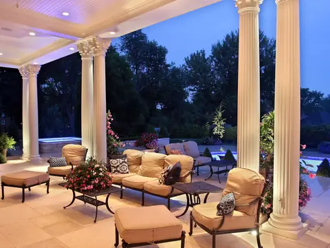 Covered columned loggia with seating and a fireplace, lit from the ceiling above, pool beyond at dusk