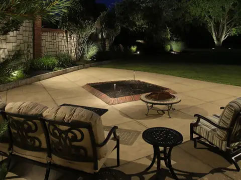 Stone terrace at night with a sunken fire feature, seating and a lit wall and planting behind