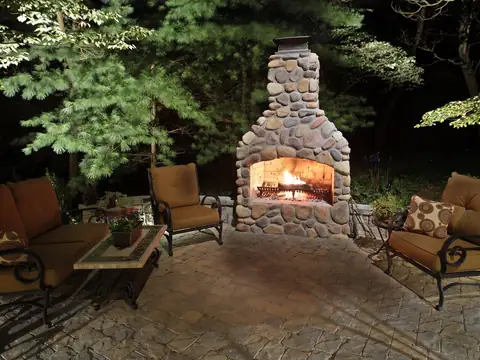 Stone outdoor fireplace lit with seating around it under mature trees