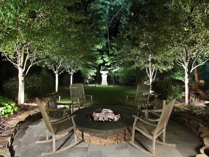 Fire pit and seating on a stone patio at night with the surrounding trees lit from within the canopy
