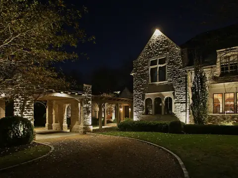 Stone and timber house at night with a lit drive curving past mature trees toward a lit outbuilding