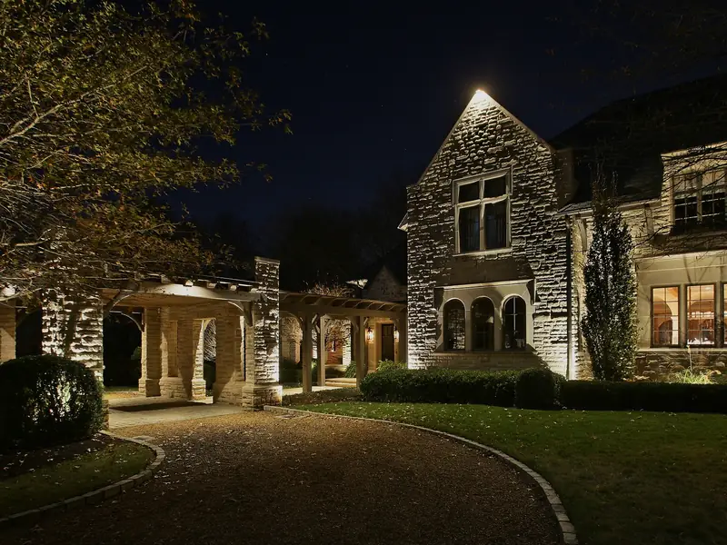Stone and timber house at night with a lit drive curving past mature trees and a lit outbuilding beyond