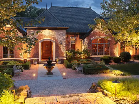 Stone home with a lit fountain in the forecourt and warm light across the gables and planting