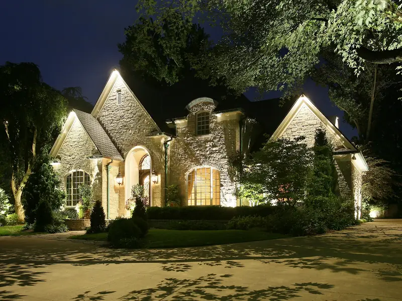 Steep-gabled stone house at night, lit through and beneath surrounding mature trees with the entry brightest