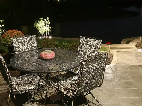 Stone patio at night with a table and chairs lit softly and the lawn and water beyond in darkness