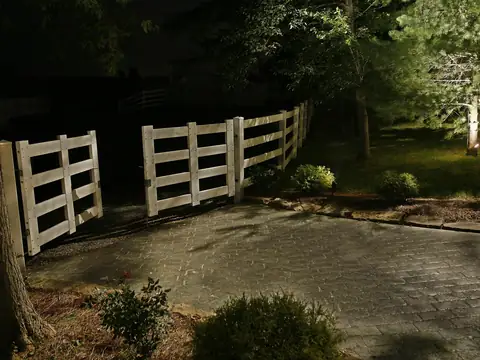 White fence and gate lit at night with a flowering tree beyond and the drive surface lit