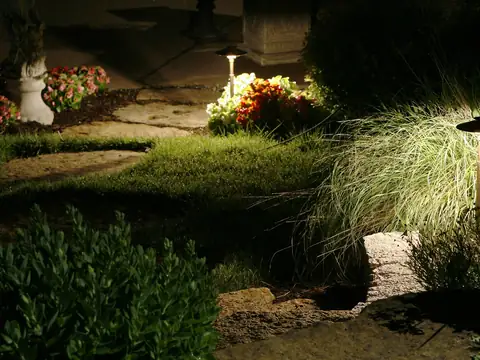Planting bed at night with a path fixture lighting ornamental grasses and a stone ornament