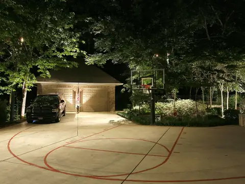 Wide paved court and basketball area lit evenly at night with trees lit at the perimeter